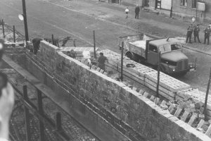 Unscharf im Vordergrund am linken Bildrand ist eine Hand mit Fotokamera zu erkennen. Quer durch das Bild zieht sich die Mauer aus Hohlblocksteinen, die gerade aufgebaut wird. Dahinter sieht man Arbeiter, eine Straßensperre aus Betonelementen, einen Lastwagen, drei Polizisten und zwei Passanten.