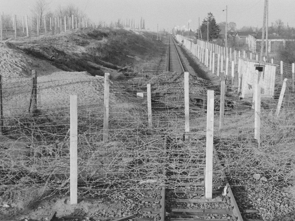 Quer über die Eisenbahnschienen verlaufen Stacheldrahtverhaue, dahinter liegt ein Haufen Schotter auf den Gleisen.