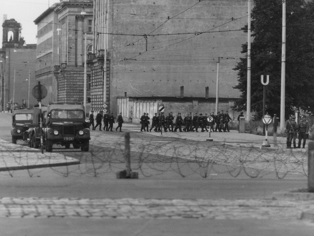Aufmarsch von Volkspolizisten an der Ebert-/Ecke Leipziger Straße, 13. August 1961 Leicht verschwommen zieht sich im Vordergrund eine Stacheldrahtrolle durchs Bild. Dahinter sind zwei Militärfahrzeuge und eine Gruppe von Volkspolizisten in der ansonsten menschenleeren Straße zu sehen.