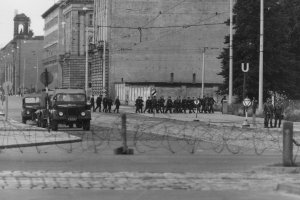 Leicht verschwommen zieht sich im Vordergrund eine Stacheldrahtrolle durchs Bild. Dahinter sind zwei Militärfahrzeuge und eine Gruppe von Volkspolizisten in der ansonsten menschenleeren Straße zu sehen.