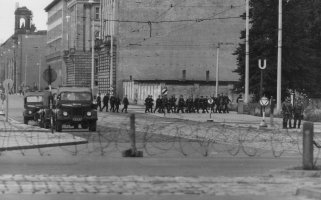 Leicht verschwommen zieht sich im Vordergrund eine Stacheldrahtrolle durchs Bild. Dahinter sind zwei Militärfahrzeuge und eine Gruppe von Volkspolizisten in der ansonsten menschenleeren Straße zu sehen.