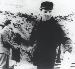 Der junge Erich Honecker steht zentral im Bild, in der rechten Hand hält er einen Hammer. Im Bildhintergrund ist ein mehrere Meter hoher Trümmerberg zu sehen, davor stehen ein Mann und eine Frau.