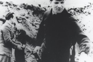 Der junge Erich Honecker steht zentral im Bild, in der rechten Hand hält er einen Hammer. Im Bildhintergrund ist ein mehrere Meter hoher Trümmerberg zu sehen, davor stehen ein Mann und eine Frau.