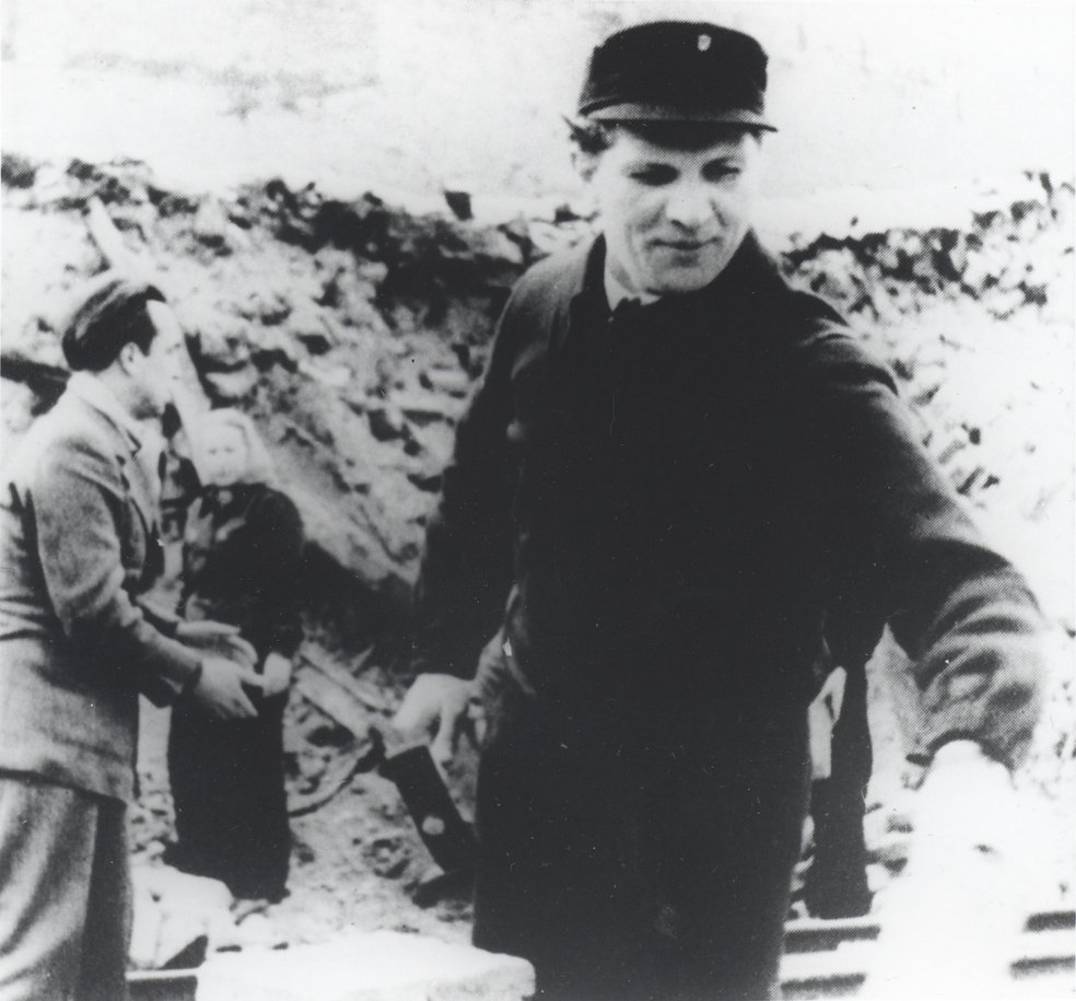 Der junge Erich Honecker steht zentral im Bild, in der rechten Hand h&auml;lt er einen Hammer. Im Bildhintergrund ist ein mehrere Meter hoher Tr&uuml;mmerberg zu sehen, davor stehen ein Mann und eine Frau.