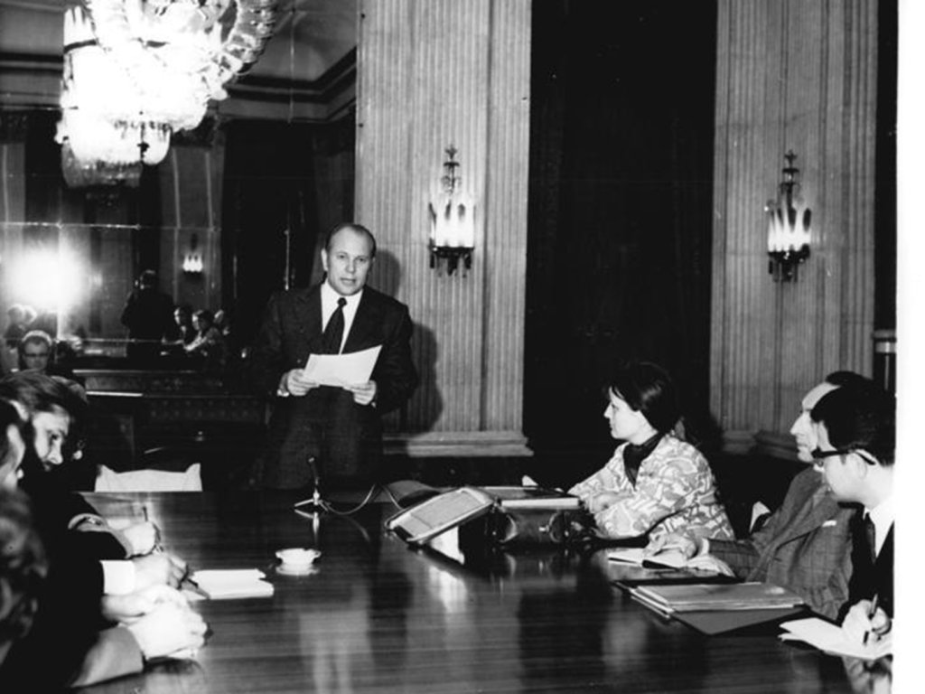 Der Botschafter steht zentral im Bild, liest von einem Blatt und blickt in die Kamera. Links und rechts von ihm sitzen Menschen an einem großen Tisch und machen Notizen.