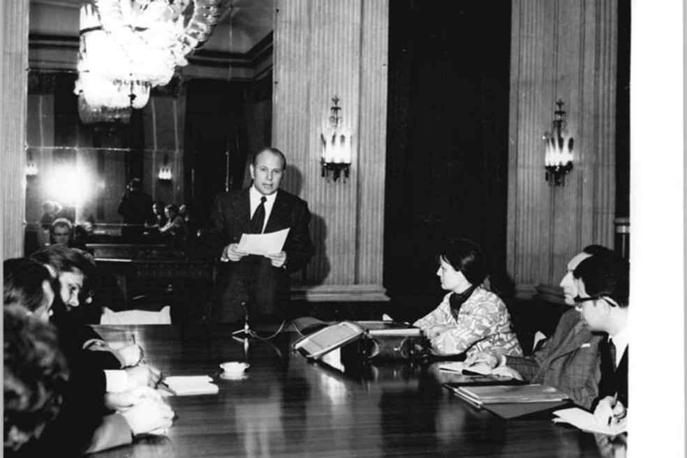 Der Botschafter steht zentral im Bild, liest von einem Blatt und blickt in die Kamera. Links und rechts von ihm sitzen Menschen an einem gro&szlig;en Tisch und machen Notizen.