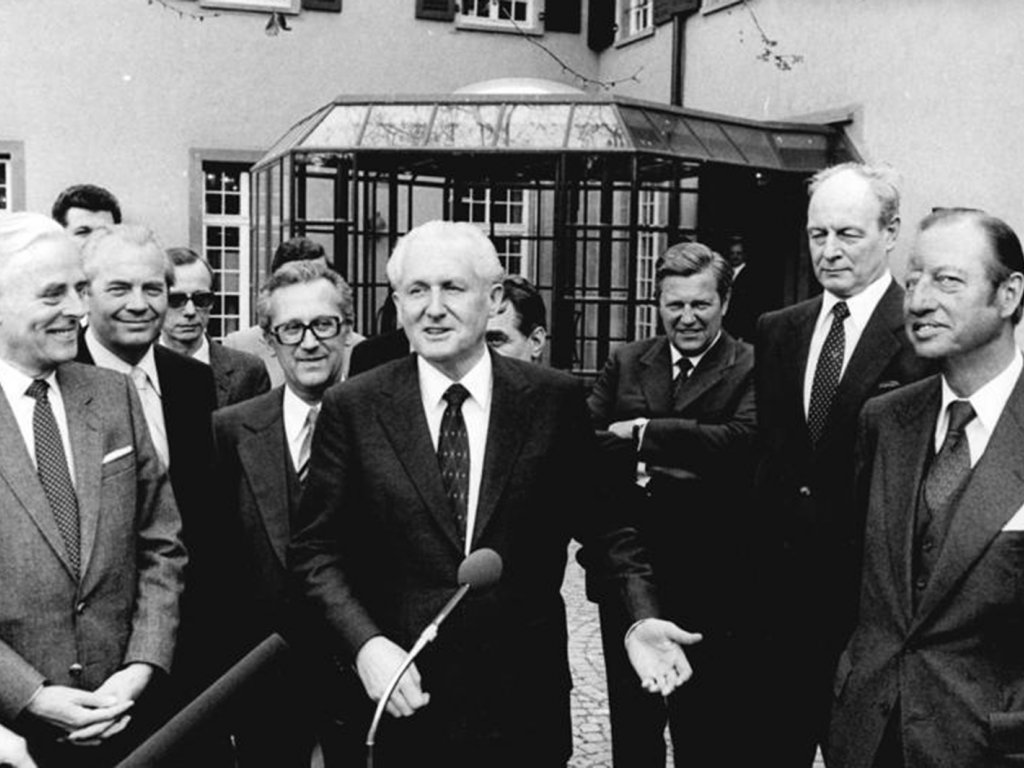ZK-Wirtschaftssekretär Günter Mittag in der Bundesrepublik mit Politikern und Industriellen der Firma Hoechst (v. l. n. r.) Willi Hoerkens, Ewald Moldt, Gerhard Beil, Günter Mittag und Rolf Sammet; Aufnahme April 1980  Günter Mittag steht im Bildzentrum, umringt von anderen, lächelnden, Männern. Er gestikuliert und spricht in ein Mikrofon.