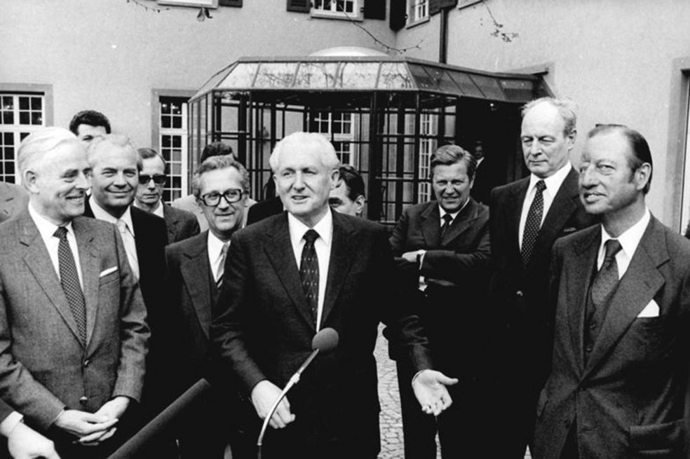 G&uuml;nter Mittag steht im Bildzentrum, umringt von anderen, l&auml;chelnden, M&auml;nnern. Er gestikuliert und spricht in ein Mikrofon.