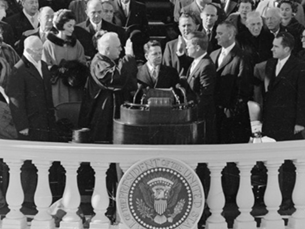 Im Vordergrund ist die weiße Brüstung des Kapitols mit dem Siegel der USA zu sehen. Kennedy steht dahinter neben einem Rednerpult gegenüber von Bundesrichter Earl Warren. Beide halten ihre rechte Hand erhoben. Sie sind von Zuschauern umgeben, links im Bild steht Jackie Kennedy.