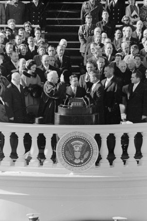 Im Vordergrund ist die weiße Brüstung des Kapitols mit dem Siegel der USA zu sehen. Kennedy steht dahinter neben einem Rednerpult gegenüber von Bundesrichter Earl Warren. Beide halten ihre rechte Hand erhoben. Sie sind von Zuschauern umgeben, links im Bild steht Jackie Kennedy.