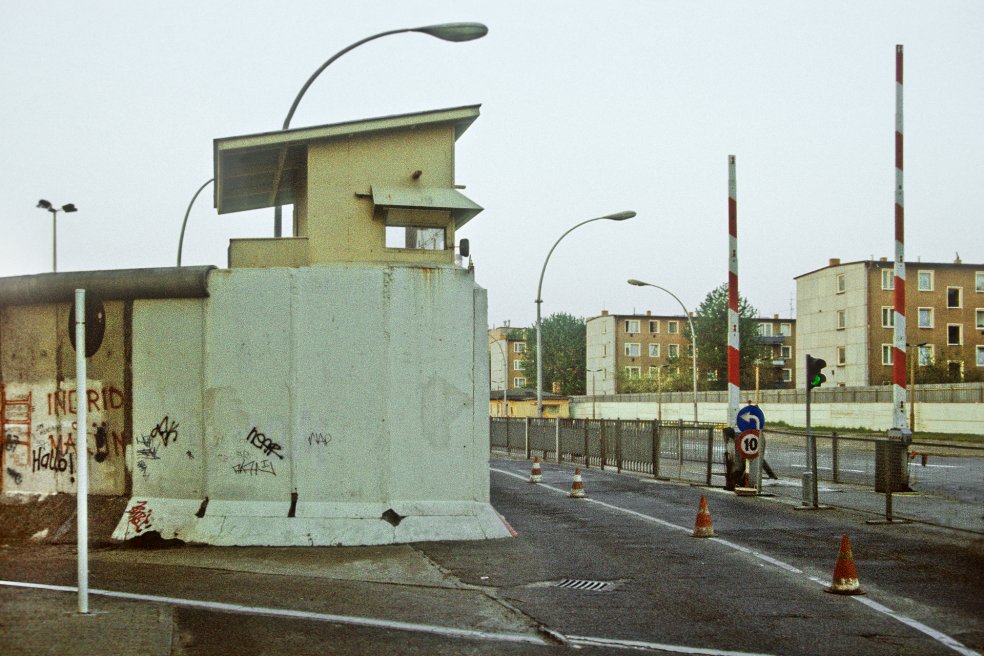 Im Vordergrund links ist ein Stück Mauer zu sehen, das mit einem Wachturm abschließt. Rechts davon befinden sich die Grenzsicherungsanlagen der Übergangsstelle Heinrich Heine Straße, dahinter stehen drei Wohnblocks.