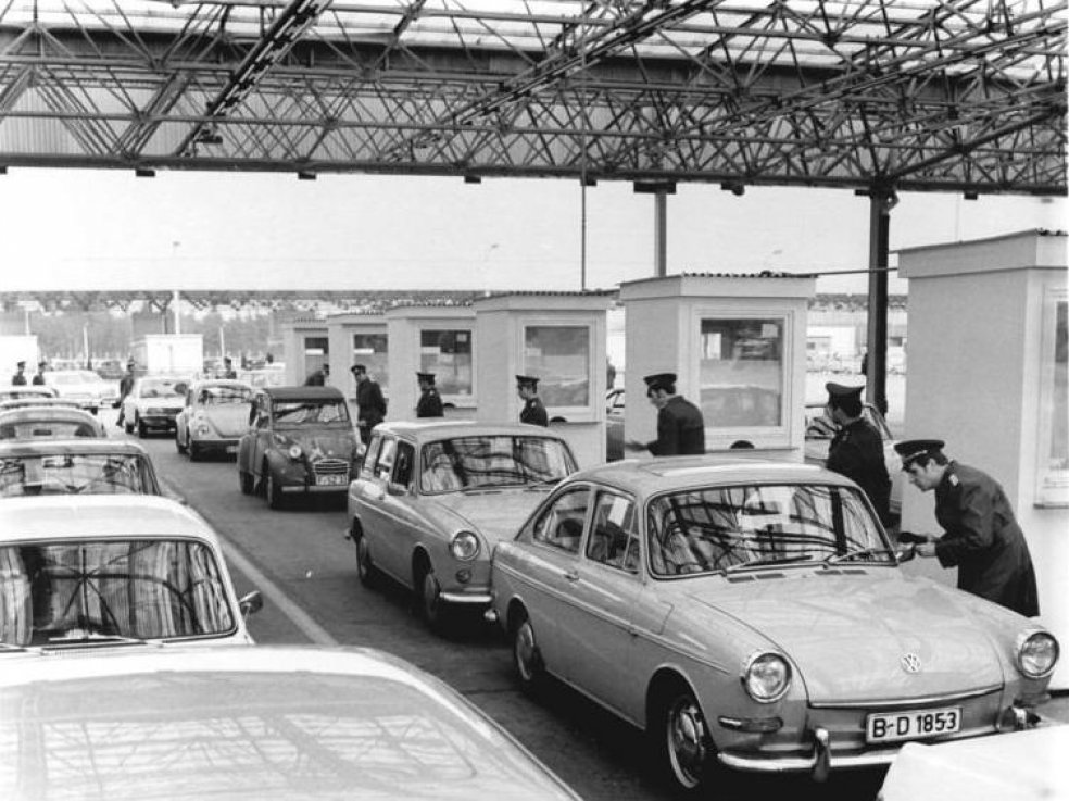 In langen Schlangen warten Autos, rechts davon ist eine Reihe Kontrollhäuschen. Daneben stehen Beamte, die die Papiere der Reisenden kontrollieren.