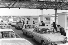 In langen Schlangen warten Autos, rechts davon ist eine Reihe Kontrollhäuschen. Daneben stehen Beamte, die die Papiere der Reisenden kontrollieren.