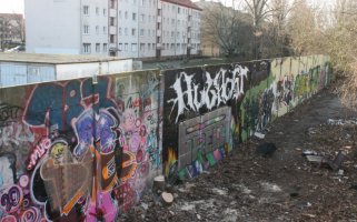 Ein mit Graffiti buntbemaltes Stück der Hinterlandmauer zieht sich von vorne links nach hinten rechts durch das Bild. Hinter der Mauer stehen Wohnblöcke. Auf der Vorderseite liegt vereinzelt Müll auf dem Erdboden.