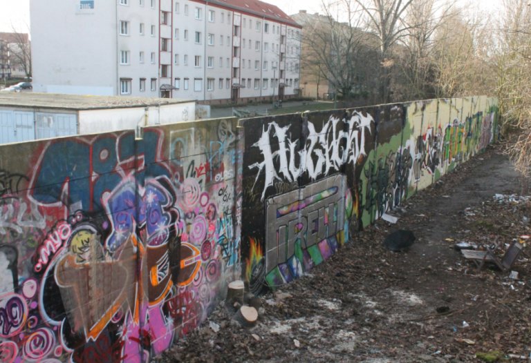 Ein mit Graffiti buntbemaltes Stück der Hinterlandmauer zieht sich von vorne links nach hinten rechts durch das Bild. Hinter der Mauer stehen Wohnblöcke. Auf der Vorderseite liegt vereinzelt Müll auf dem Erdboden.
