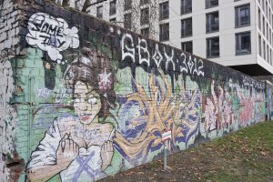 Mit Graffiti bemalte Hohlblocksteinmauer. Am hinteren Ende wird die Mauer überragt von einem neu errichten Gebäude.