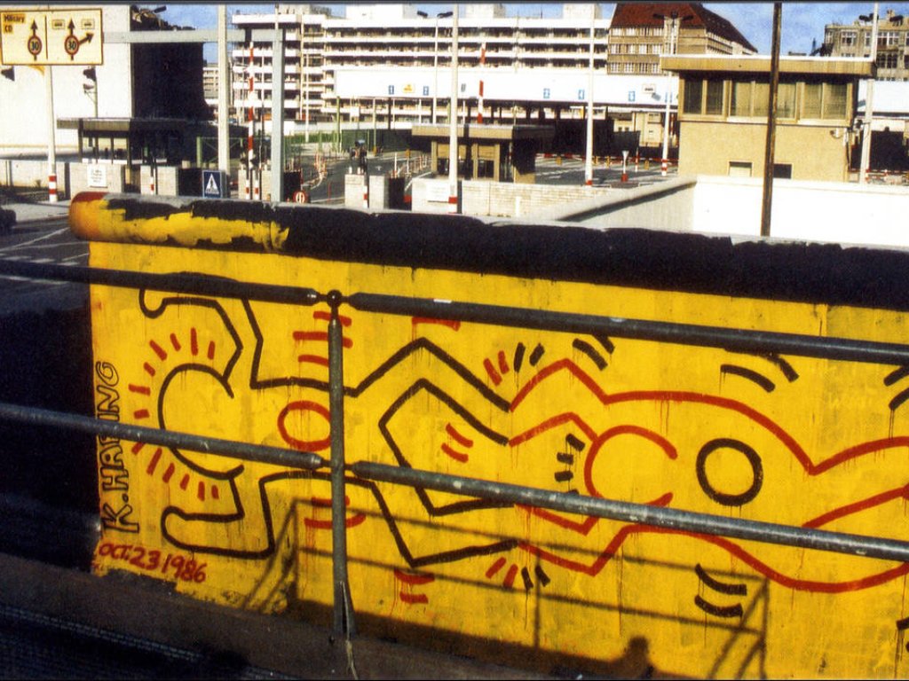 Im Zentrum ist ein Stück Mauer direkt am Grenzübergang. Darauf ist eine leuchtend gelbe Fläche mit zwei von Harings Figuren, die Horizontal gemalt sind. Eine Figur hält die andere an den Füßen. Am Mauerrand steht: K. Haring. Oft. 23 1986