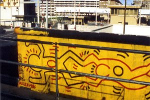 Im Zentrum ist ein St&uuml;ck Mauer direkt am Grenz&uuml;bergang. Darauf ist eine leuchtend gelbe Fl&auml;che mit zwei von Harings Figuren, die Horizontal gemalt sind. Eine Figur h&auml;lt die andere an den F&uuml;&szlig;en. Am Mauerrand steht: K. Haring. Oft. 23 1986