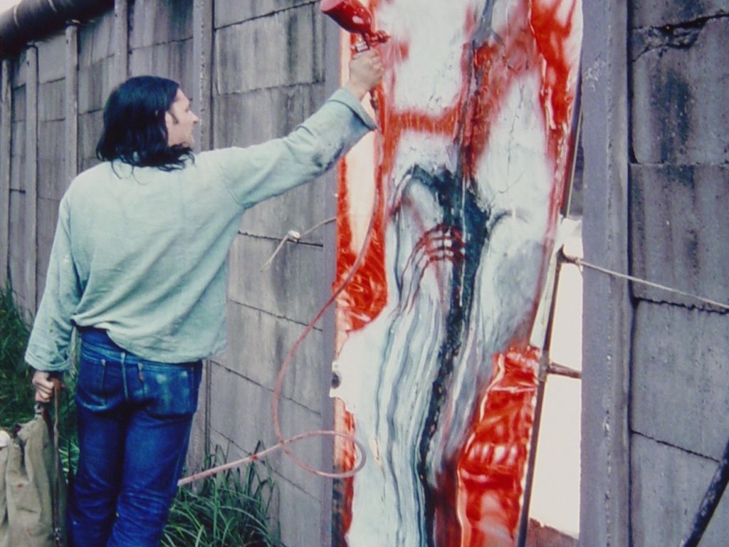 Elsner steht auf einem Haufen herausgebrochener Mauerzeile. Neben ihm in der Mauerlücke ist sein abstraktes Bild eingesetzt, das er mit einer Sprühpistole mit roter Farbe weiter bearbeitet.
