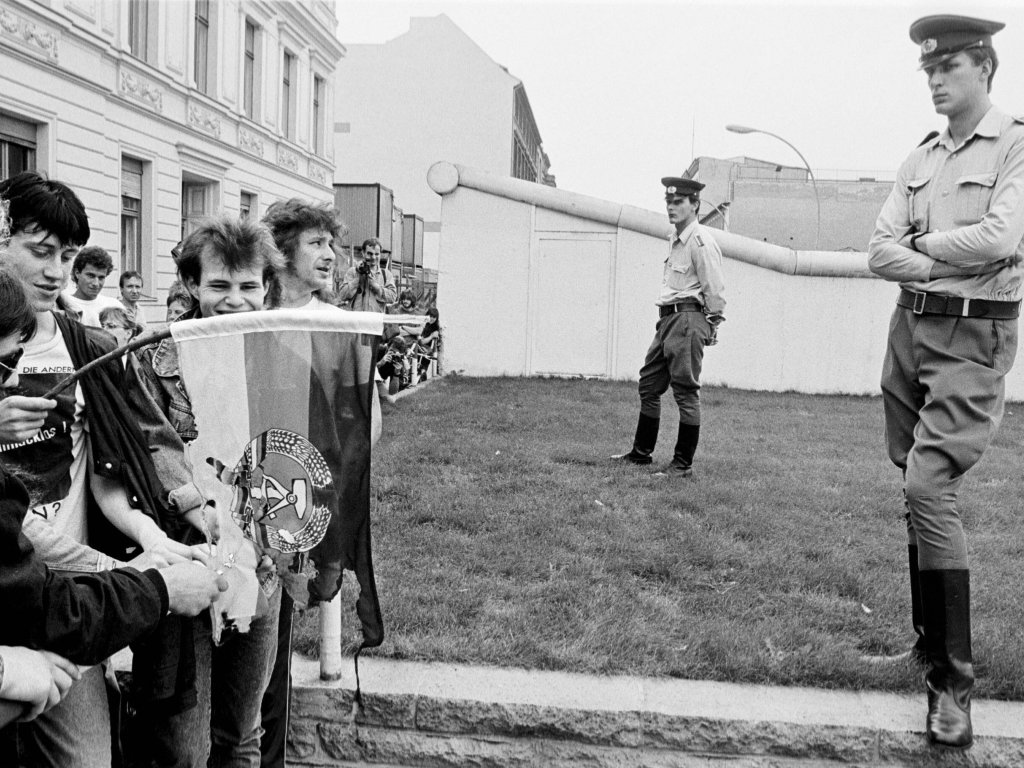 1989: Protestaktion zum 28. Jahrestag des Mauerbaus. West-Berliner Demonstranten verbrennen vor den Grenzsoldaten am Checkpoint Charlie eine DDR-Flagge.