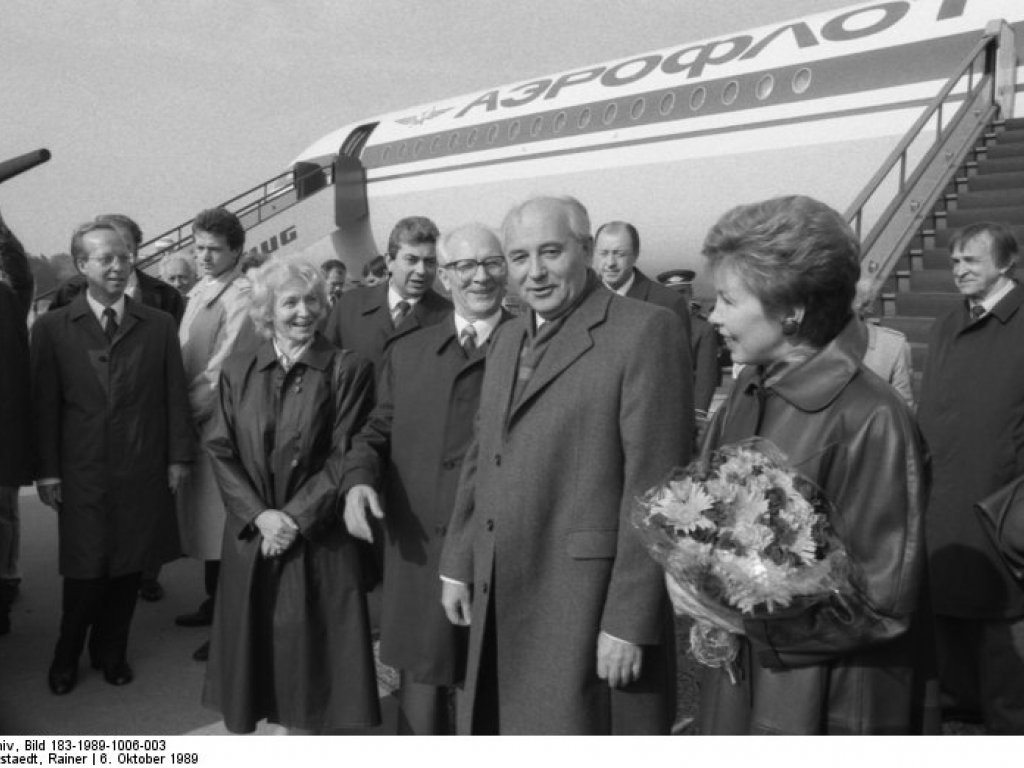 Im Vordergrund sind der lächelnde Michail Gorbatschow und seine Frau Raisa zu sehen, die einen Blumenstrauß hält. Links hinter ihnen, ebenfalls lächelnd, Margot und Erich Honecker. Weitere Menschen umringen die Gruppe, ein Mann hält ein Mikrofon. Hinter Ihnen steht ein Aeroflot Flugzeug mit angestellter Treppe.