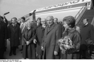 Im Vordergrund sind der lächelnde Michail Gorbatschow und seine Frau Raisa zu sehen, die einen Blumenstrauß hält. Links hinter ihnen, ebenfalls lächelnd, Margot und Erich Honecker. Weitere Menschen umringen die Gruppe, ein Mann hält ein Mikrofon. Hinter Ihnen steht ein Aeroflot Flugzeug mit angestellter Treppe.
