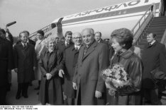 Im Vordergrund sind der lächelnde Michail Gorbatschow und seine Frau Raisa zu sehen, die einen Blumenstrauß hält. Links hinter ihnen, ebenfalls lächelnd, Margot und Erich Honecker. Weitere Menschen umringen die Gruppe, ein Mann hält ein Mikrofon. Hinter Ihnen steht ein Aeroflot Flugzeug mit angestellter Treppe.