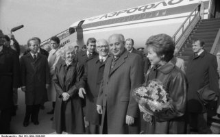 Im Vordergrund sind der lächelnde Michail Gorbatschow und seine Frau Raisa zu sehen, die einen Blumenstrauß hält. Links hinter ihnen, ebenfalls lächelnd, Margot und Erich Honecker. Weitere Menschen umringen die Gruppe, ein Mann hält ein Mikrofon. Hinter Ihnen steht ein Aeroflot Flugzeug mit angestellter Treppe.