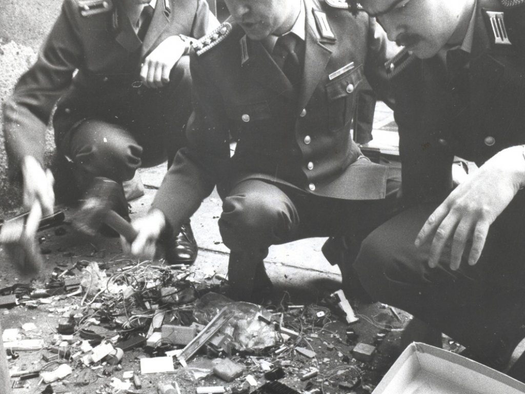 Drei uniformierte Männer der Volkspolizei hocken auf dem Boden. Die zwei Männer links schlagen mit Hämmern auf einen vor ihnen liegenden Haufen elektrotechnischer Teile ein.