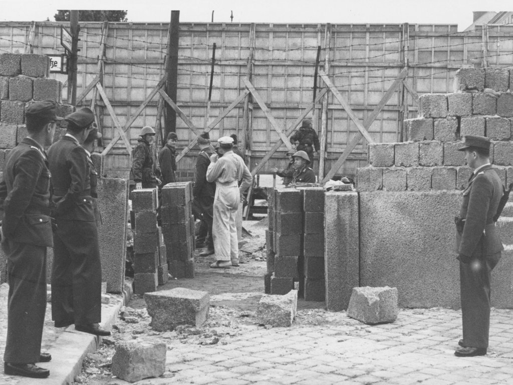 Durch die Bildmitte geht eine Mauer aus Hohlblocksteinen, in der eine große Lücke klafft. Im Vordergrund stehen Polizisten und schauen durch die Mauerlücke auf eine Gruppe Soldaten und Arbeiter, die vor einer mehrere Meter hohen Absperrung aus Holz und Stacheldraht stehen.