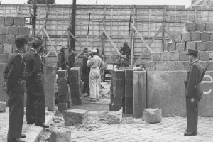 Durch die Bildmitte geht eine Mauer aus Hohlblocksteinen, in der eine große Lücke klafft. Im Vordergrund stehen Polizisten und schauen durch die Mauerlücke auf eine Gruppe Soldaten und Arbeiter, die vor einer mehrere Meter hohen Absperrung aus Holz und Stacheldraht stehen.