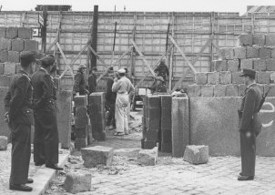 Durch die Bildmitte geht eine Mauer aus Hohlblocksteinen, in der eine große Lücke klafft. Im Vordergrund stehen Polizisten und schauen durch die Mauerlücke auf eine Gruppe Soldaten und Arbeiter, die vor einer mehrere Meter hohen Absperrung aus Holz und Stacheldraht stehen.