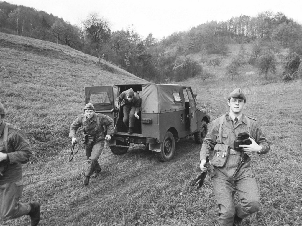 Aus der hinteren Tür eines Militärfahrzeuges springt ein bewaffneter Soldat. Drei weitere laufen im Vordergrund auf die Kamera zu.