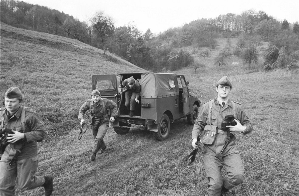 Aus der hinteren Tür eines Militärfahrzeuges springt ein bewaffneter Soldat. Drei weitere laufen im Vordergrund auf die Kamera zu.