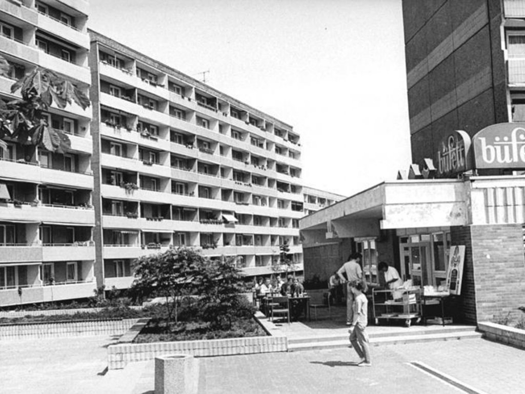 Die RAF-Terroristin Susanne Albrecht wohnte von 1980 bis Anfang 1985 in der DDR im Neubaugebiet Sachsendorf-Madlow in Cottbus; Aufnahme 12. Juni 1990  Dominant im Bild links steht ein Plattenbau mit Balkonen. Es ist sommerliches Wetter. Auf der rechten Seite stehen und sitzen einige Menschen auf der Terrasse eines Lokals, auf dessen Dach die Beschriftung büffett angebracht ist. Dahinter erhebt sich ein weiterer Plattenbau.