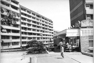 Dominant im Bild links steht ein Plattenbau mit Balkonen. Es ist sommerliches Wetter. Auf der rechten Seite stehen und sitzen einige Menschen auf der Terrasse eines Lokals, auf dessen Dach die Beschriftung büffett angebracht ist. Dahinter erhebt sich ein weiterer Plattenbau.