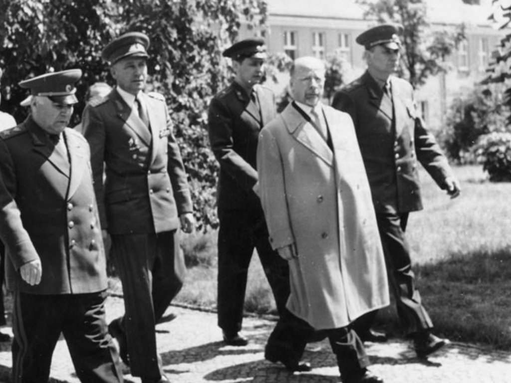 Walter Ulbricht geht in der Bildmitte und blickt in die Kamera. Neben und hinter ihm geht eine Gruppe uniformierter Männer.