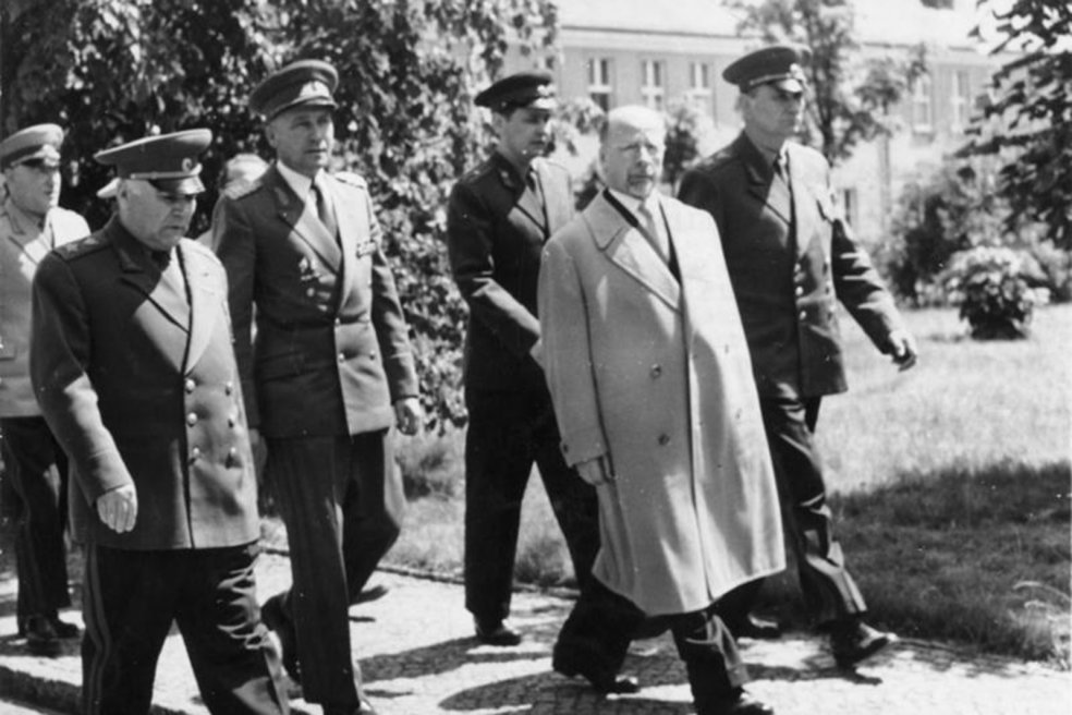 Walter Ulbricht geht in der Bildmitte und blickt in die Kamera. Neben und hinter ihm geht eine Gruppe uniformierter M&auml;nner.