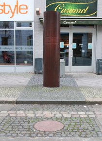 Die bronzefarbene Gedenksäule für Peter Fechter. Davor ist ein Pflastersteinstreifen zu sehen, der den ehemaligen Verlauf der Berliner Mauer markiert und eine runde, in den Boden eingelassene Granitplatte.
