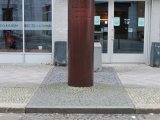 Die bronzefarbene Gedenksäule für Peter Fechter. Davor ist ein Pflastersteinstreifen zu sehen, der den ehemaligen Verlauf der Berliner Mauer markiert und eine runde, in den Boden eingelassene Granitplatte.