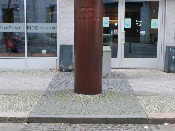 Gedenkstele Peter Fechter Die bronzefarbene Gedenksäule für Peter Fechter. Davor ist ein Pflastersteinstreifen zu sehen, der den ehemaligen Verlauf der Berliner Mauer markiert und eine runde, in den Boden eingelassene Granitplatte.