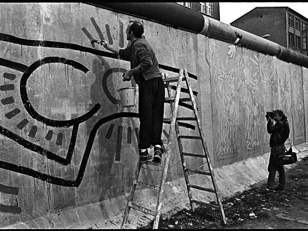 Keith Haring steht auf einer Leiter und ist dabei, seine Figuren zu malen. Rechts im Bild steht ein Mann und fotografiert ihn.