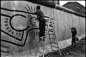 Keith Haring steht auf einer Leiter und ist dabei, seine Figuren zu malen. Rechts im Bild steht ein Mann und fotografiert ihn.