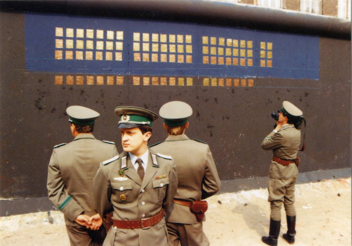 Vor der schwarz-blau grundierten, teilweise mit Kupferplatten beklebten Mauer stehen vier Grenzsoldaten. Drei stehen in einer Gruppe im Vordergrund, die Rücken einander zugewandt. Ein vierter filmt die Installation mit einer Kamera. Vor der schwarz-blau grundierten, teilweise mit Kupferplatten beklebten Mauer stehen vier Grenzsoldaten. Drei stehen in einer Gruppe im Vordergrund, die Rücken einander zugewandt. Ein vierter filmt die Installation mit einer Kamera.