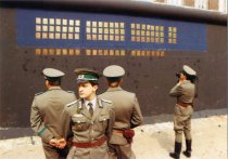 Vor der schwarz-blau grundierten, teilweise mit Kupferplatten beklebten Mauer stehen vier Grenzsoldaten. Drei stehen in einer Gruppe im Vordergrund, die Rücken einander zugewandt. Ein vierter filmt die Installation mit einer Kamera.