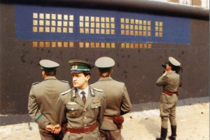 Vor der schwarz-blau grundierten, teilweise mit Kupferplatten beklebten Mauer stehen vier Grenzsoldaten. Drei stehen in einer Gruppe im Vordergrund, die Rücken einander zugewandt. Ein vierter filmt die Installation mit einer Kamera.