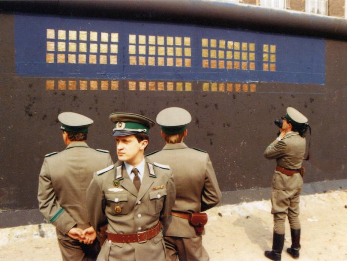 Vor der schwarz-blau grundierten, teilweise mit Kupferplatten beklebten Mauer stehen vier Grenzsoldaten. Drei stehen in einer Gruppe im Vordergrund, die Rücken einander zugewandt. Ein vierter filmt die Installation mit einer Kamera.